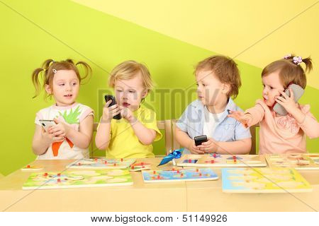 Two boys and two girls with phones in their hands are sitting at a table with toys