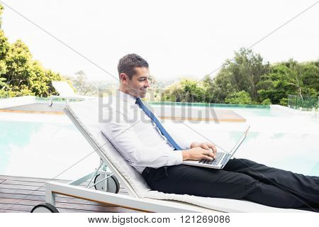 Smarty dressed using laptop near poolside while relaxing on sun lounger