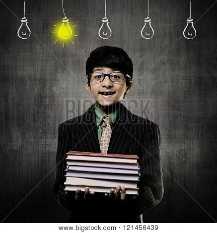 Cute Intelligent Little Boy Holding Books And Wearing Glasses Smiling While Standing Before A Chalkboard 5 Bulbs of ideas hanging and one bulb is on