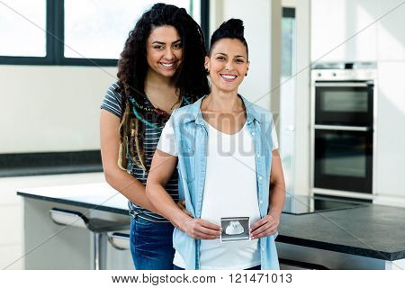 Pregnant lesbian couple holding a sonography report and smiling in kitchen