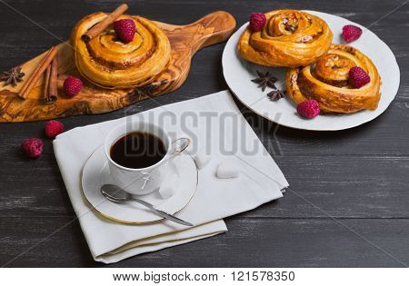 Round twisted Denish Buns with berries raspberries syrup white cup of espresso coffee sugar cubes cinnamon sticks star anise cutting board made of wood olive on a black wooden background