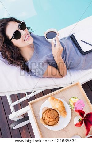 Portrait of young woman having cup of tea and relaxing on a sun lounger near poolside