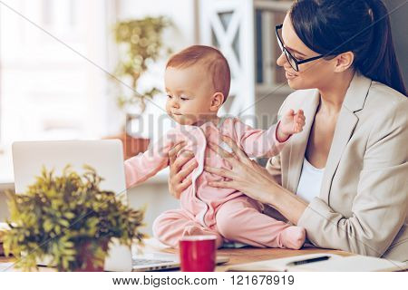 Working together with joy. Cheerful young beautiful businesswoman looking at her baby girl with smile while sitting at her working place
