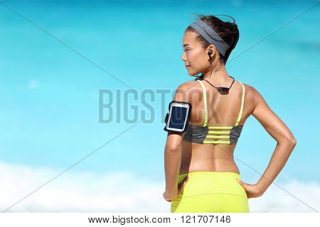 Fitness runner woman with fit back wearing phone armband and wireless headphones on ocean background. Multiracial exercise girl jogging on summer beach with sports smartphone touchscreen for app.