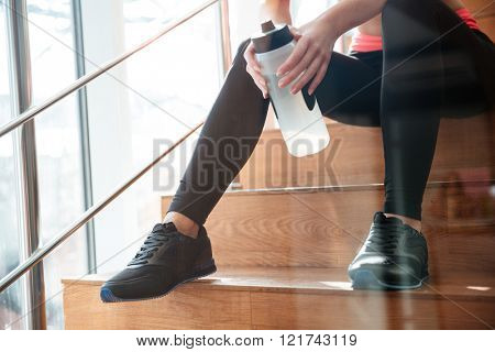 Beautiful slim legs of attractive woman athlete sitting on stairs in gym with bottle of water
