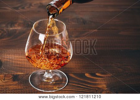 Pouring Brandy Or Cognac From The Bottle Into The Glass Against Wooden Background