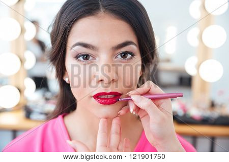 Hands of female makeup artist with lip brush applying red lipstick to young attrative woman lips in beauty salon