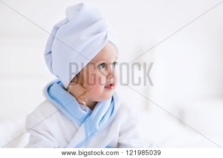 Child after bath. Cute little girl with wet curly hair wearing a bathrobe and head towel sitting on a white bed using lotion and brush. Hygiene for kids. Bathroom textile for babies and children.