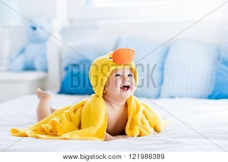 Happy laughing baby wearing yellow hooded duck towel sitting on parents bed after bath or shower. Clean dry child in bedroom. Bathing and washing of little kids. Children hygiene. Textile for infants.