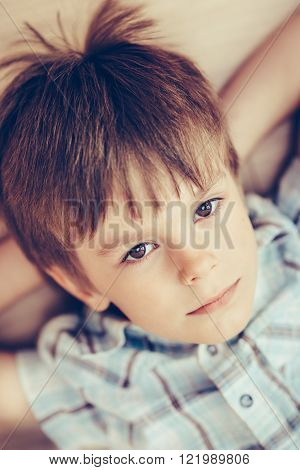 Closeup portrait of pensive little boy with brown eyes wearing checkered shirt lying on floor and looking at camera.