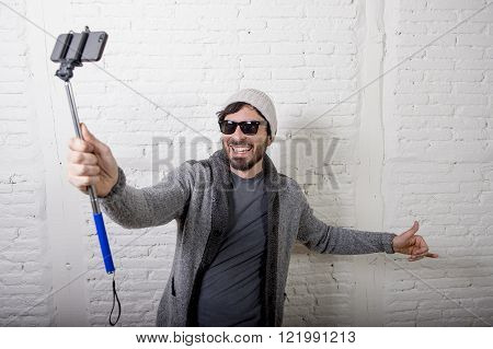 young attractive man in casual clothes beanie hipster style holding selfie stick mobile phone shooting self portrait photo or recording video in internet blog and blogger concept