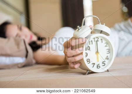 Asian beautiful woman sleep on the bed and wake up to turn off white alarm clock at 6 am. in her bedroom.  Condo room lifestyle for working woman. Get up to go to work.