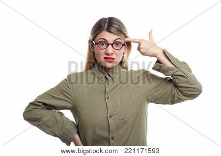 Stressed young female employee wearing red lipstick, stylish eyeglasses and khaki shirt grimacing and holding finger at her temple like a pistol as if going to shoot herself, fed up with work