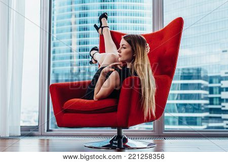 Sexy female model with fair hair posing on stylish red armchair against panoramic window with view of skyscrapers.