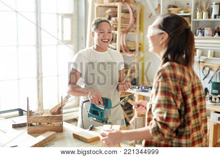 Laughing carpenter with powertool looking at her colleague during talk in workshop