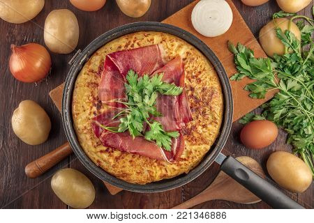 An overhead photo of a Spanish tortilla in the traditional frying pan, with jamon and parsley, on dark rustic textures with ingredients