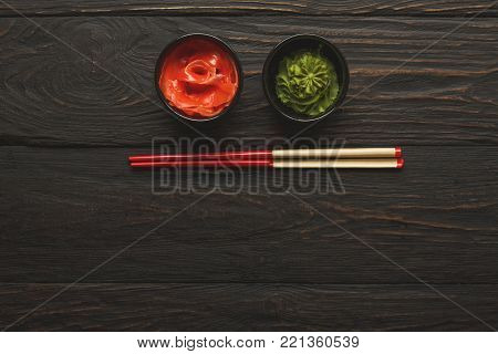 Pieces of traditional japanese tablewear. Red chopsticks with hierogliphs, small black bowls with ginger and wasabi on dark wooden table. Top view, flat lay, copy space, background