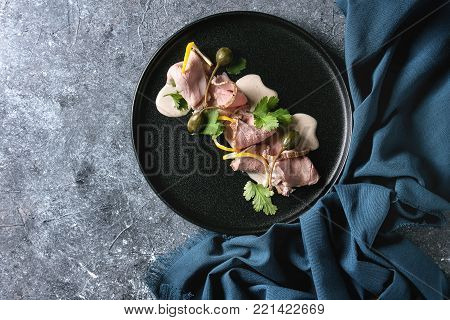 Vitello tonnato italian dish. Thin sliced veal with tuna sauce, capers and coriander served on black plate with blue textile over blue texture background. Top view, copy space