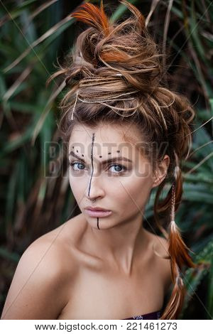 Portrait of attractive serios girl with painting on her face and high bunch of hair. There are black line and dots on her face. There are pigtails and feathers in her hair. She is looking at camera
