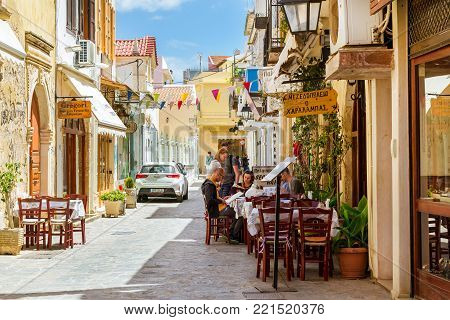 Rethymno, Greece - May 3, 2016: Tourists read menu at tables in street cafe with national cuisine. Street advertising signboard on touristic street. Mediterranean architecture and cuisine on Crete