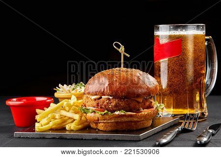 Tasty burger, french fries with sauce and glass of beer on black board. Still life. Served at restaurant, cafe, bar, pub cheesburger, hamburger
