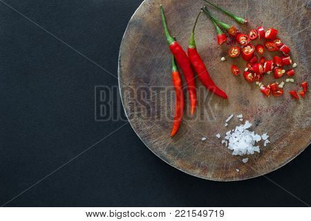 Red hot chili peppers and sliced peppers on a wooden cutting board with salt crystals nearby on a black background. Asian species.