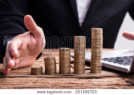 Close-up Of A Stacked Golden Coins And Businessperson's Hand On Wooden Desk