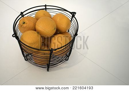 Many bright Mandarin oranges fill up a black metal basket on a white surface and background.