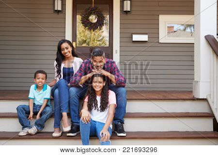 Family With Children Sit On Steps Leading Up To Porch Of Home