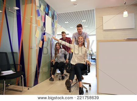 Young cheerful business people in smart casual wear having fun while racing on office chairs and smiling