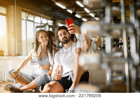 Young attractive smiling active fitness couple sitting on the floor of the gym and taking a selfie, leaning on the mirror with a towel.
