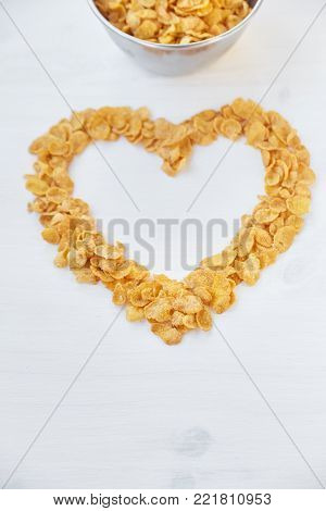 The symbol of the heart is laid out of cornflakes. Cornflakes in a metal bowl on a painted white wooden background.