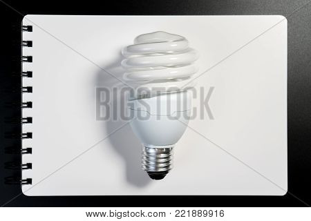 One fluorescent light bulb on white notebook paper on black background for energy saving concept. Light bulb on white book for business and education thinking idea concept.