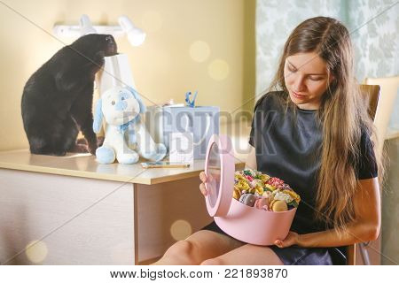 Beautiful girl with flowers in a box on his birthday. The gifts on the table. Black cat looks in the package