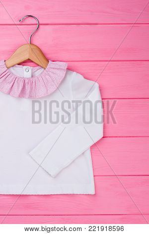 Adorable blouse for baby girl on wooden hanger. White top decorated with pink ruffle collar. Kids fashion.