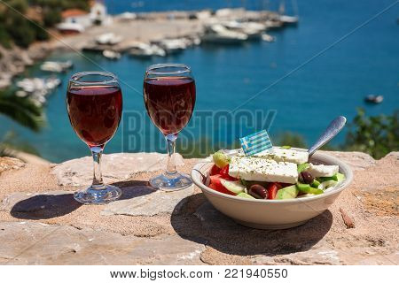 Two glasses of red wine and bowl of greek salad with greek flag on a stone terrace by the sea view, summer greek holidays concept. Horizontal. Daylight.