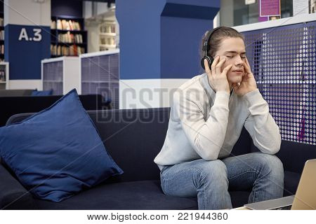 Handsome university student in sweater and jeans using wireless headset while listening to information, preparing for test in college. Cute guy enjoying music in headphones, having dreamy look