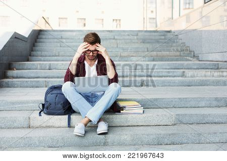 Desperate student sitting on stairs and working on laptop outdoors in university campus. Man raised his hands to his head. Technology, education and remote working concept, copy space