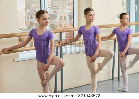 Three Young Ballerinas Posing At Ballet Barre. Beautiful Ballet Dancers Practicing Dance Move In Stu