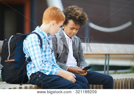 Two Boys With Bacpacks Behind Their Backs Sitting In The City And Using Mobile Phone Together After 