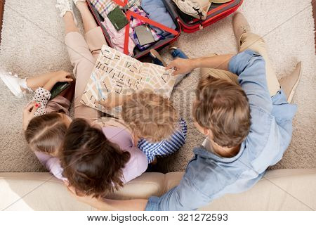 Overview of young family of parents, their son and daughter looking at map guide while going for journey or camping