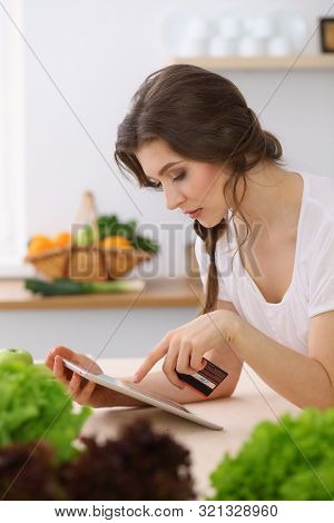 Young Woman Looking For A New Recipe For Cooking In A Kitchen. Housewife Is Making Online Shopping B