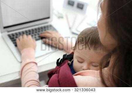 Young woman with baby in ergo carrier using laptop indoors