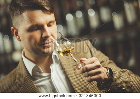 Sommelier Smelling Flavor Of Whiskey In Bokal On Background Of Shelves With Bottles In Cellar.