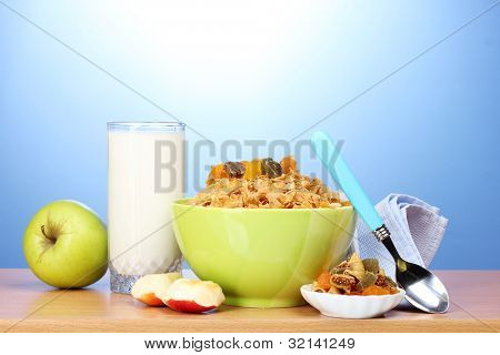 leckere Cornflakes in grüner Schale, Äpfel und Glas Milch auf Holztisch auf blauem Hintergrund