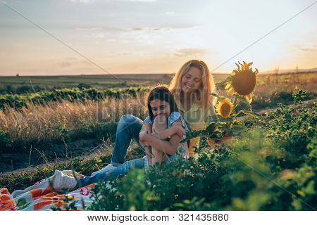 Teen Brunette Girl Sitting Calmy At The Plaid Among The Mealow And Hugging Her Legs, Her Young Mom I