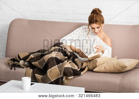 Attractive Woman In White Sweater Sitting On Sofa And Reading Book