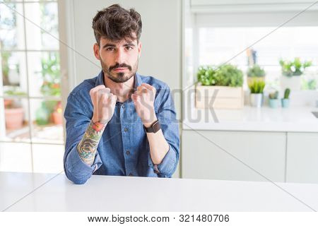 Young man wearing casual shirt sitting on white table Ready to fight with fist defense gesture, angry and upset face, afraid of problem