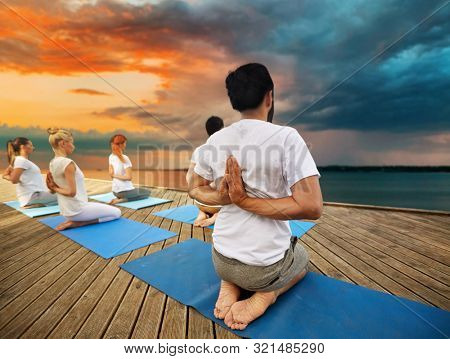 fitness, sport, yoga and healthy lifestyle concept - group of people exercising in reverse prayer pose on sea pier over sunset background