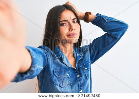 Beautiful woman wearing denim shirt make selfie by camera over isolated white background confuse and wondering about question. Uncertain with doubt, thinking with hand on head. Pensive concept.
